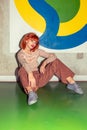 Young sylish redhead Caucasian woman in retro style of 70s sitting at wall vertical looking into camera Royalty Free Stock Photo