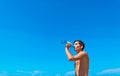 Young tanned smiling guy drinks water from a bottle against a blue cloudless sky