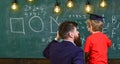 Young teacher explaining arithmetic to the little boy in academic cap. Man with beard and blond child turned back in the