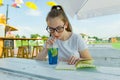 Young teen girl with glasses drinking blue lemonade and reading book in an outdoor cafe. Background green area of rest and enterta Royalty Free Stock Photo