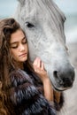 A young teenage girl in a fur coat, with long curly hair in a soft blue dress hugs a white horse on the Bank of the oken Royalty Free Stock Photo