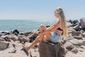 A young teenage girl with long blonde hair blonde sits on the rocks of the breakwater near the sea shore on the beach Royalty Free Stock Photo