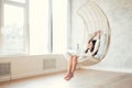 Young teenage girl relaxing in comfortable hanging chair near window at home. Child sitting in chair and chilling out. Relax conce