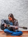 Teenager brunette girl with long hair sitting on the floor and playing an black acoustic guitar on gray wall background Royalty Free Stock Photo