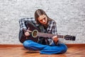 Teenager brunette girl with long hair sitting on the floor and playing an black acoustic guitar on gray wall background Royalty Free Stock Photo