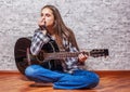 Teenager brunette girl with long hair sitting on the floor and playing an black acoustic guitar on gray wall background Royalty Free Stock Photo