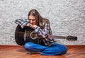 Teenager brunette girl with long hair sitting on the floor and playing an black acoustic guitar on gray wall background Royalty Free Stock Photo