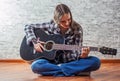 Teenager brunette girl with long hair sitting on the floor and playing an black acoustic guitar on gray wall background Royalty Free Stock Photo