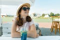 Young teenager girl in hat sunglasses smiling and drinking cool fruit cocktail on a hot summer day in outdoor cafe Royalty Free Stock Photo