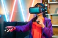 Young teenager girl playing video game on virtural reality or VR headset using joystick at home on neon background -