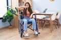 Young teenager girl sitting on wheelchair at the living room with angry face, negative sign showing dislike with thumbs down, Royalty Free Stock Photo