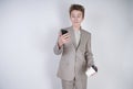 Young teenager with surprised emotions in grey business clothes standing with mobile phone and paper cup of coffee on white studio