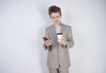 Young teenager with surprised emotions in grey business clothes standing with mobile phone and paper cup of coffee on white studio