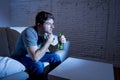 Young television addict man sitting on home sofa watching TV and drinking beer bottle