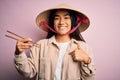 Young thai woman wearing traditional conical asian hat eating food using chopsticks with surprise face pointing finger to himself