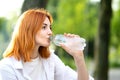 Young thirsty redhead woman drinking water from a bottle in summer park