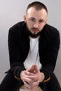 Young thoughtful bearded man with short dark hair in black jacket, white T-shirt, sitting at stool, interlacing fingers. Royalty Free Stock Photo