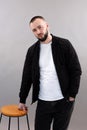 Young thoughtful bearded man with short dark hair wearing black jacket, jeans, white T-shirt, standing at wooden stool. Royalty Free Stock Photo