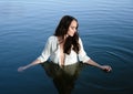 Young thoughtful brunette woman in white shirt is standing in the lake and moving hands through water. Royalty Free Stock Photo