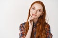 Young thoughtful ginger girl in shirt, without makeup, looking at camera and thinking, making choice, standing over