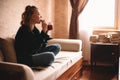 Young woman drinking tea and eating chocolate while looking through window sitting on sofa in living room at home Royalty Free Stock Photo