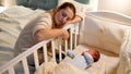 Young tired and exhausted mother fallen asleep while rocking crib of her newborn baby at night. Concept of sleepless