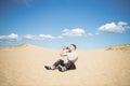 Young tired man drinking water while sitting on the top of sand dune in desert, sand is al around and some greens. Hot summer weat Royalty Free Stock Photo