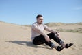 Young tired man sitting on the top of sand dune in desert, sand is al around and some greens. Hot summer weather, concept of trave Royalty Free Stock Photo