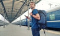 Young tourist backpacker with smartphone stand on railway station platform and waiting for train. Travel concept. Copy space