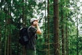 Young tourist in comfortable clothes and with a backpack stands in a mountain forest on a background of beautiful coniferous trees