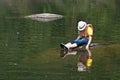 Young tourist girl sitting on stone in middle of lake. Bowing head in hat thoughtfully and keeping fingers in water Royalty Free Stock Photo