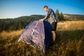 Young tourist man setting up tent on autumn meadow, traveling high in mountains sunset