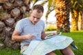 Young tourist sitting under palm tree with mobile phone and tablet. He is looking at the map. Positive emotion. Vacation time Royalty Free Stock Photo