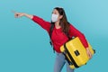 Young tourist wearing a protective mask believes COVID-19 is carrying a suitcase and points to an empty space on a blue background