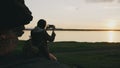 Young tourist woman backpacker photographing landscape on her smartphone camera after hiking on rock at sunset