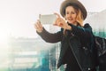 Young tourist woman in hat and with backpack stands at airport and takes photos, shoots video on smartphone`s camera. Royalty Free Stock Photo