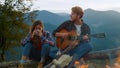 Young tourists enjoy campfire in mountains. Hipsters couple play music on nature