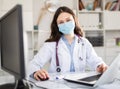 Young trainee doctor in a protective mask sits at a desktop in front of a computer in office