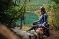 Young travel woman siting on deadwood near amazing lake on coniferous forest background. Concept of mountains trip. Side view.