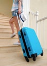 A young travel woman in white sneakers carries a blue suitcase up the stairs to the hotel