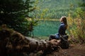 Young travel women siting on deadwood near amazing lake on coniferous forest background. Concept of mountains trip. Side view.