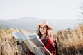 Young traveler woman wearing a hat, a red poncho and a backpack, checking the map during trek to explore nature.