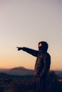 young traveler wrapped up on top of a mountain pointing finger at the sunset with beautiful mountains in the background