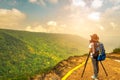 Young traveling woman with backpack hat and camera on tripod stand on the top of the mountain cliff watching beautiful view