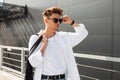 Young trendy hipster man with a stylish hairstyle straightens black sunglasses. Handsome modern guy in fashionable white clothes Royalty Free Stock Photo