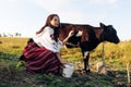 Young Ukrainian woman sits near calf with water can dressed in traditional national clothes on pasture