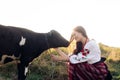 Young Ukrainian woman sits, smiles and holds calf dressed in traditional national clothes on pasture