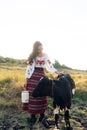 Young Ukrainian woman stands near calf with water can dressed in traditional national clothes on pasture