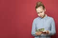 Young unhappy puzzled girl holding one us dollars cash money on pink background