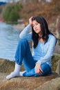 Young unhappy teen girl sitting on rocks along lake shore, looking off to side, head in hand